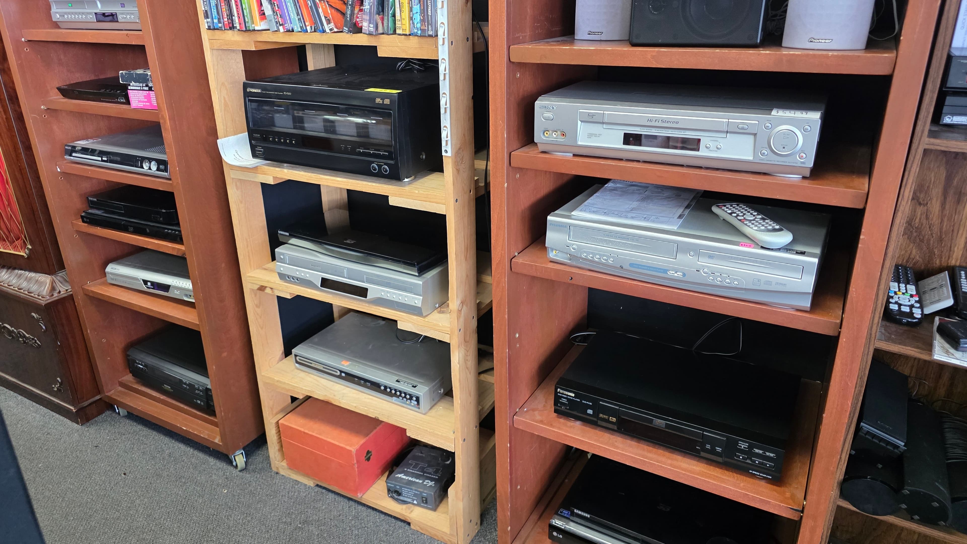 Wooden shelving racked with DVD players, VCRs and a Sony Hi-Fi VCR, Pioneer bookshelf speakers on top and remotes alongside