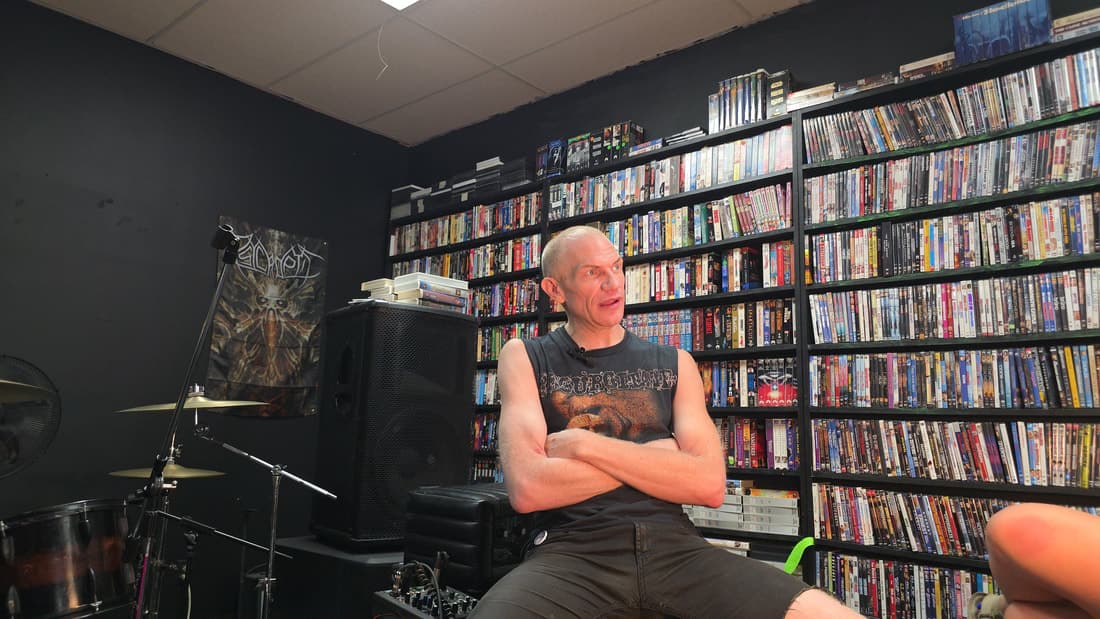 Cameron Novack seated with arms crossed during the same interview, wider framing with the drum kit visible