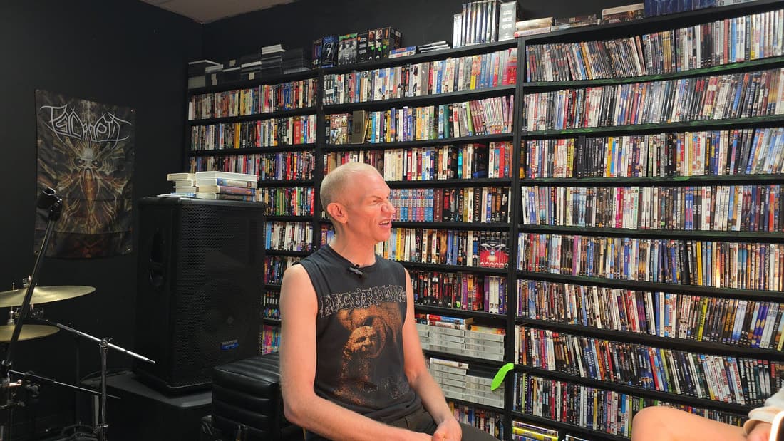 Cameron Novack seated with a clip-on lav mic, mid-sentence, against the DVD wall with a drum kit at frame left