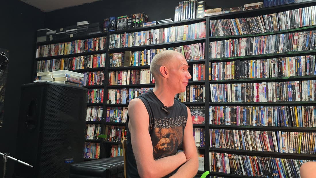 Cameron Novack seated in front of the store's DVD wall during a filmed interview, a clip-on mic at his collar.