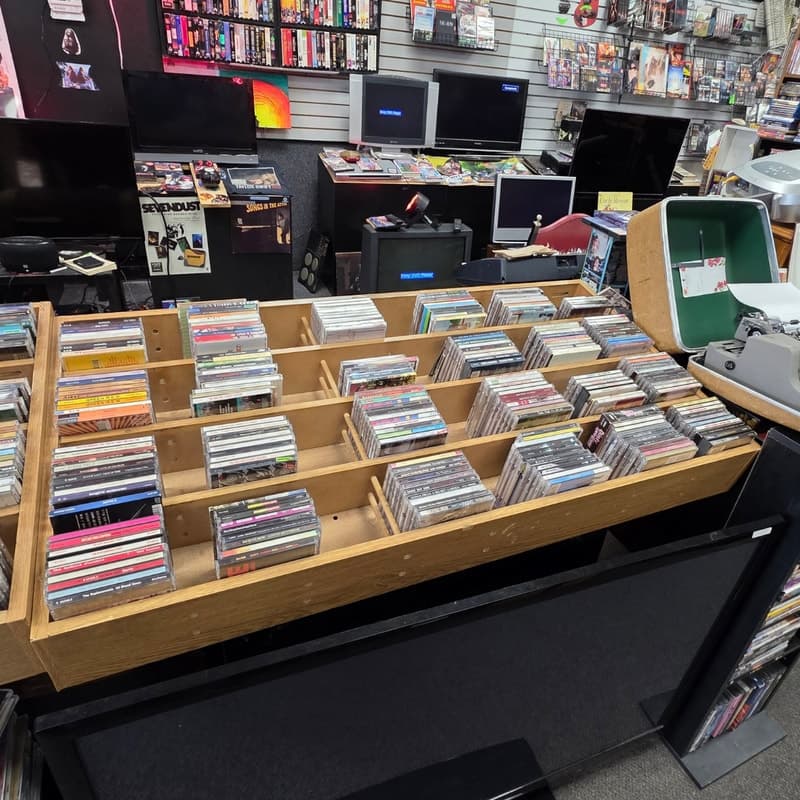 Rows of CD browser bins on a wood-topped counter with CRT TVs and the VHS wall behind