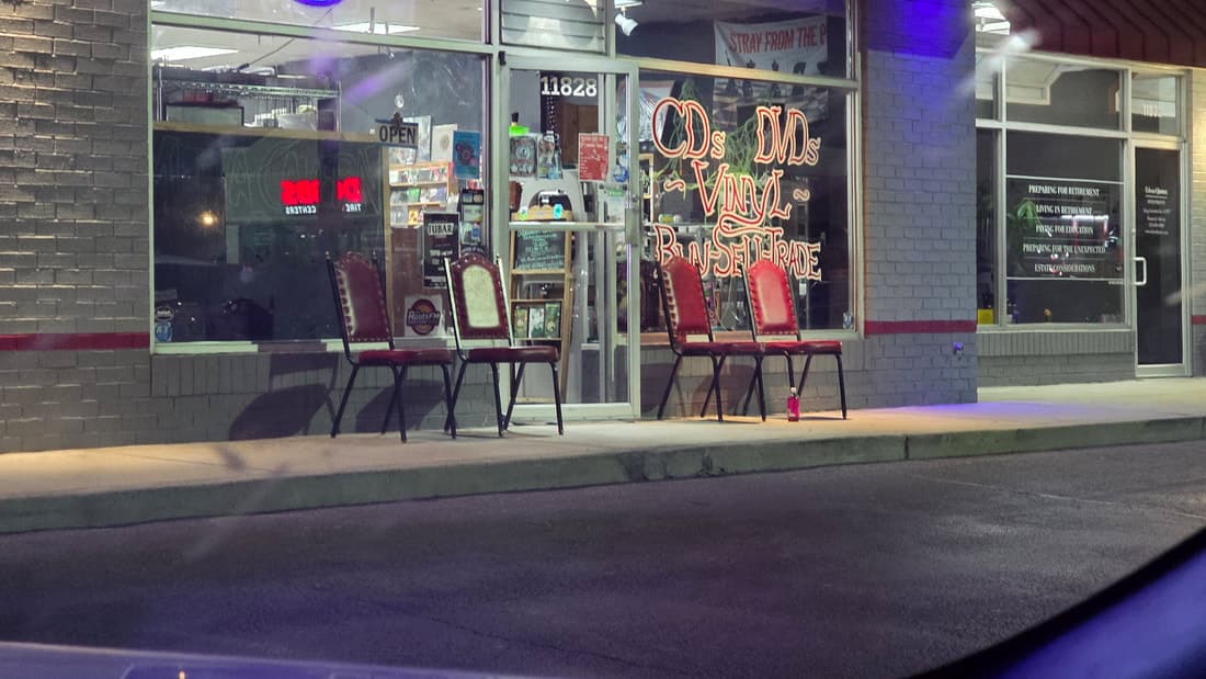 CD Warehouse storefront at night with neon window signage for CDs, DVDs and vinyl and chairs out front