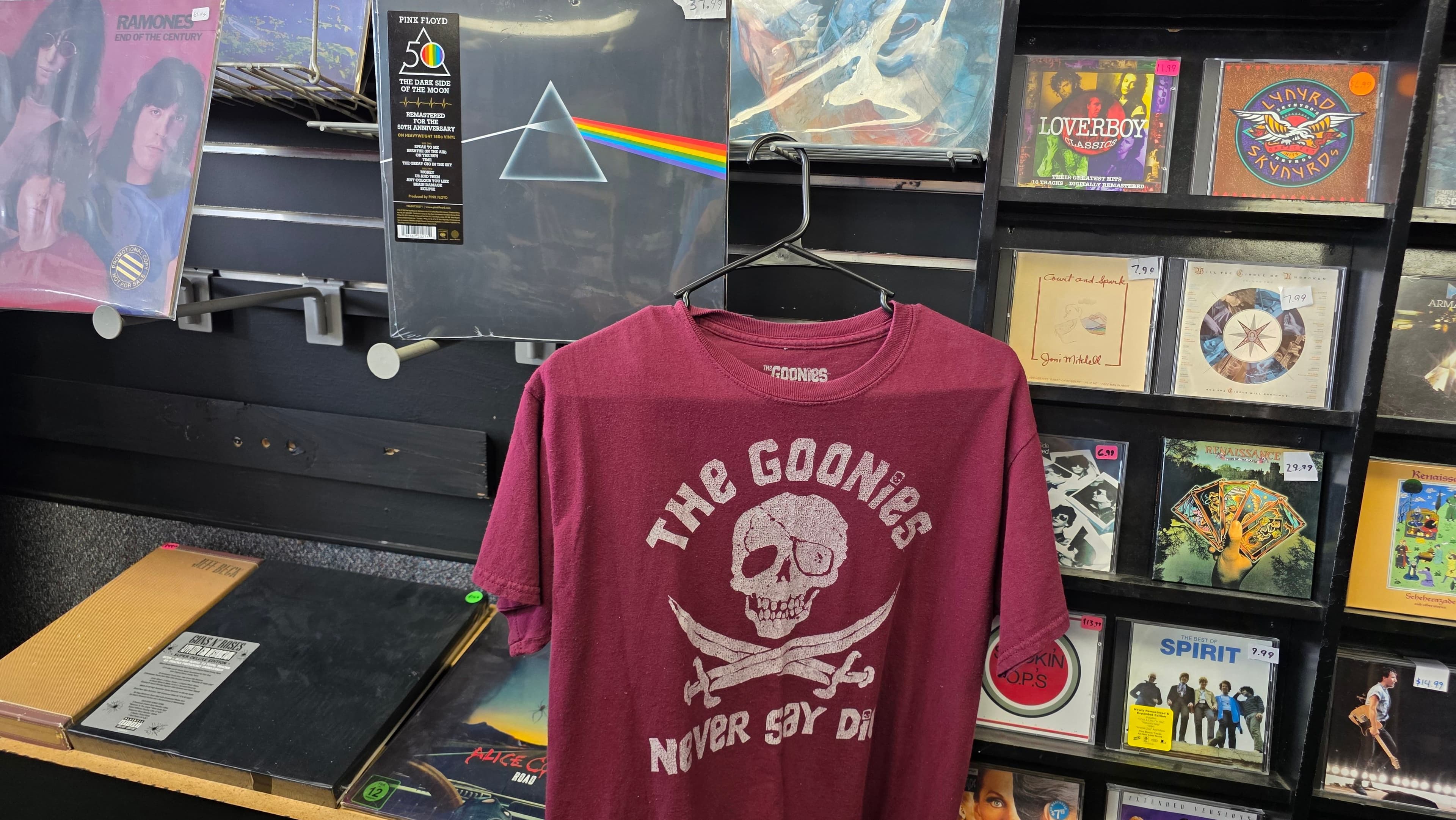 A maroon Goonies 'Never Say Die' tee on a hanger in front of the vinyl and CD wall.