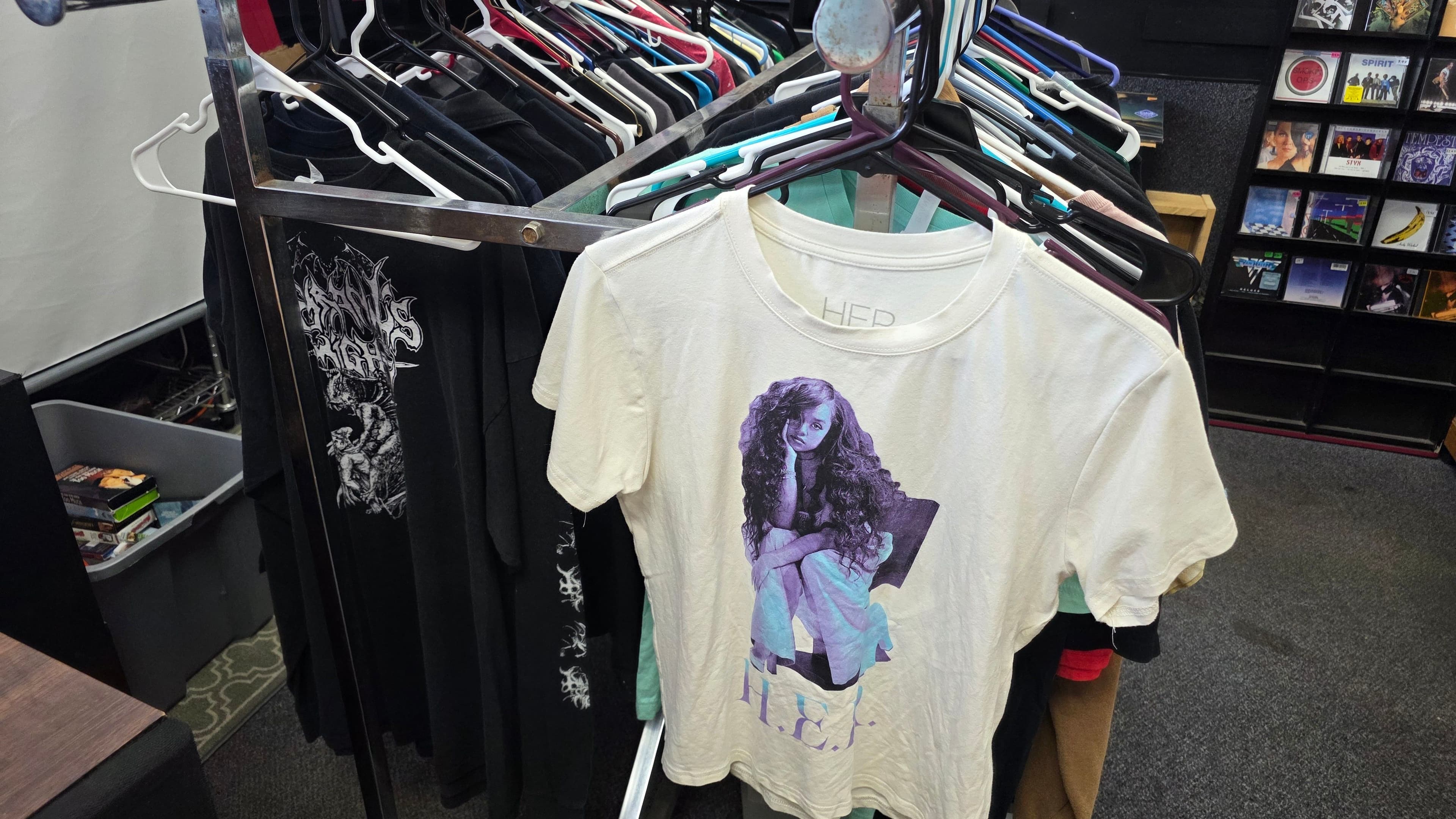 A cream H.E.R. tee hanging at the front of a packed clothing rack, black band tees behind it and CD racks beyond