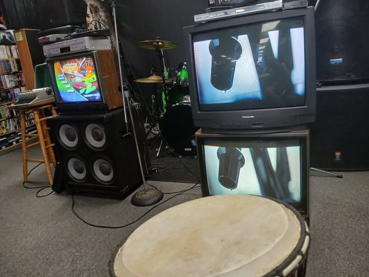 CRT screens showing a microphone close-up, with a djembe, drum kit and PA cabinet in the stage area