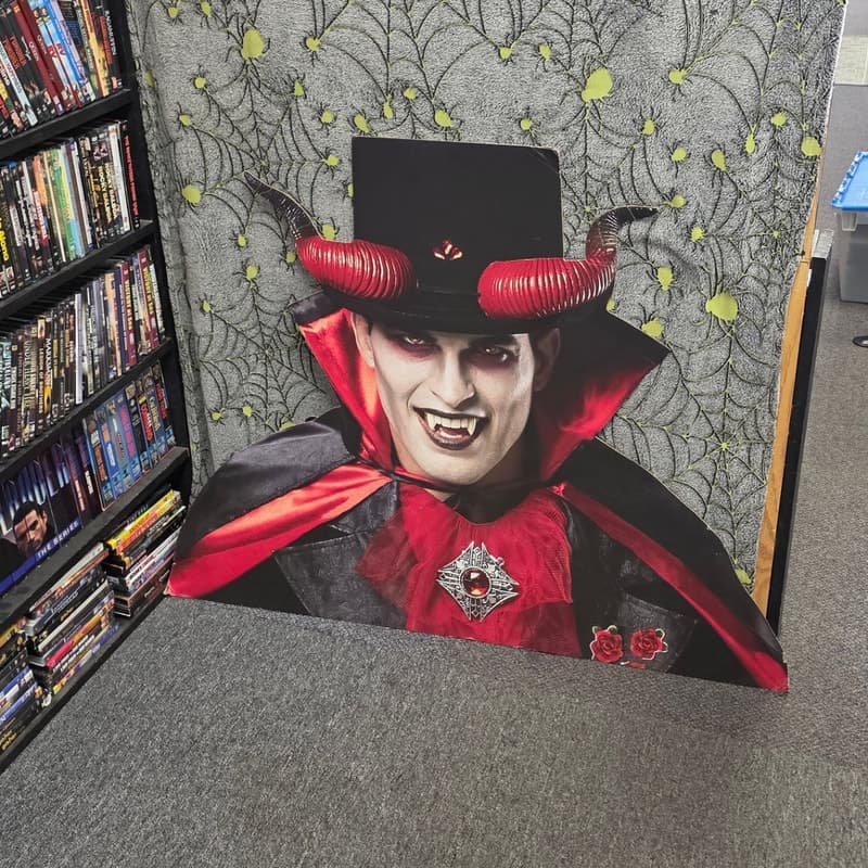 Life-size vampire cardboard standee beside DVD shelving with cobweb decor